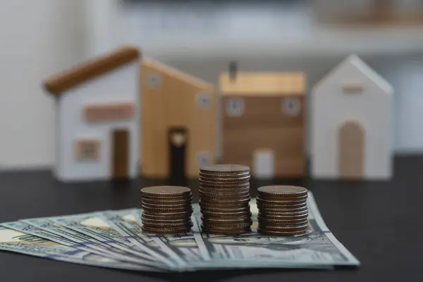 Real Estate Investment and Savings Concept. Cash and coin stacks in front of wooden house models symbolizing real estate investment.