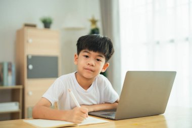 Genç bir çocuk elinde dizüstü bilgisayar ve kalemle bir masada oturuyor. Defterine bir şeyler yazmaya odaklanmış.