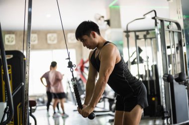 Siyah kolsuz tişörtlü ve şortlu odaklanmış bir adam modern bir spor salonunda kablo makinesiyle triseps itme egzersizi yapıyor. Biçimi ve kas yapısı iyi aydınlatılmış ortamda vurgulanmış..