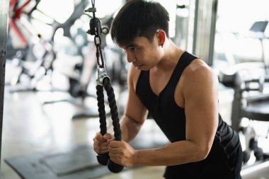 Siyah kolsuz tişörtlü ve şortlu odaklanmış bir adam modern bir spor salonunda kablo makinesiyle triseps itme egzersizi yapıyor. Biçimi ve kas yapısı iyi aydınlatılmış ortamda vurgulanmış..