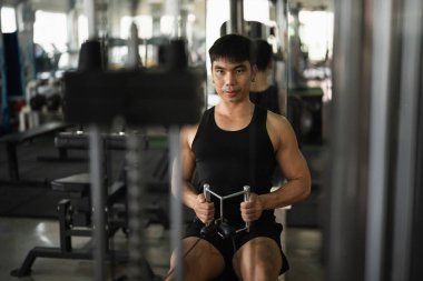 Siyah kolsuz tişörtlü bir adam modern bir spor salonundaki bir sıra makinesinde çalışıyor. Ciddi ifadesi ve kas yapısı kendini spora ve dayanıklılık eğitimine adadığını vurguluyor..