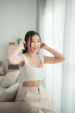 Uzun siyah saçlı neşeli genç bir kadın, beyaz bir bluz ve bej pantolon giyiyor, kulaklıklarını düzeltirken parlak bir şekilde gülümsüyor. Parlak, modern bir oturma odasında, mutluluk ve enerji saçıyor..