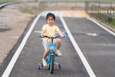 Asyalı kız bebek gülümseyerek parkta bisiklet sürmeyi öğreniyor. Çocuk bisiklet pratiği için eğitim konsepti, bebek sporları konsepti.