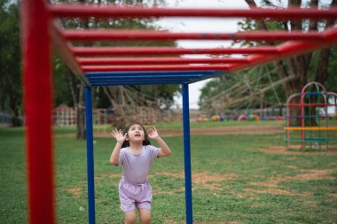 Asyalı küçük kız bir parkta kırmızı ve mavi bir yapıya bakıyor. Mor bir gömlek ve şort giyiyor.