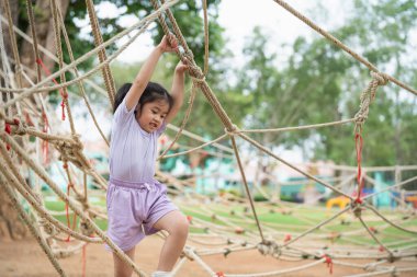 Asyalı genç bir kız halat kursunda oynuyor, iplere tırmanıp iniyor. Sahne hareketli ve eğlenceli, kız pistte gezinirken eğleniyor.