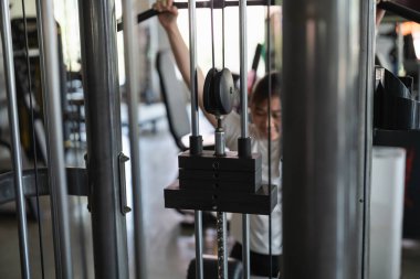 Genç bir adam bir ağırlık makinesinde çalışıyor. Ağırlıkları kaldırırken gülümsüyor.