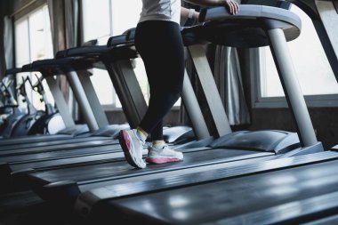 Bir kadın spor salonunda koşu bandında koşuyor. Beyaz spor ayakkabı ve siyah pantolon giyiyor.