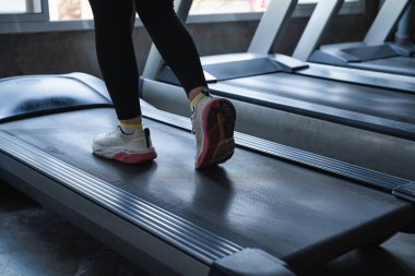Bir kadın koşu bandında koşuyor. O, siyah pantolon ve beyaz ayakkabılar giyiyor. Kadın spor yaparken azim ve odaklanma kavramı