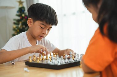 İki çocuk satranç oynuyor. Biri gülümsüyor ve diğeri kaşlarını çatıyor. Beyaz gömlekli çocuk gülümsüyor ve turuncu gömlekli kız kaşlarını çatıyor.