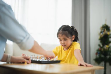 Asyalı genç kız bir yetişkinle satranç oynuyor. Kadın kızı oynarken izliyor ve muhtemelen ona oyunun nasıl oynanacağını öğretiyor. Kız sarı bir gömlek giyiyor.