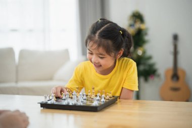 Asyalı genç kız bir yetişkinle satranç oynuyor. Kadın kızı oynarken izliyor ve muhtemelen ona oyunun nasıl oynanacağını öğretiyor. Kız sarı bir gömlek giyiyor.