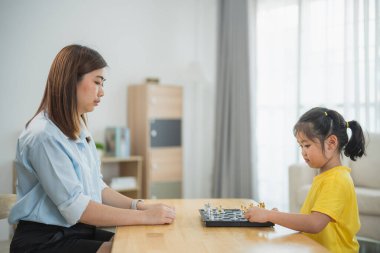 Asyalı genç kız bir yetişkinle satranç oynuyor. Kadın kızı oynarken izliyor ve muhtemelen ona oyunun nasıl oynanacağını öğretiyor. Kız sarı bir gömlek giyiyor.