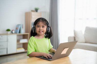 Genç bir kız önünde dizüstü bilgisayarla bir masada oturuyor. Kulaklık takıyor ve gülümsüyor. Oda kitaplarla dolu ve duvarda bir saat var.