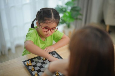 Asyalı bir kadın ve bir çocuk satranç oynuyorlar. Kadın gözlük takıyor ve çocuk yeşil bir gömlek giyiyor. Sahne neşeli ve neşeli.