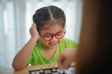 Gözlüklü Asyalı genç kız bir yetişkinle satranç oynuyor. Kız gülümsüyor ve oyundan hoşlanıyor gibi görünüyor.