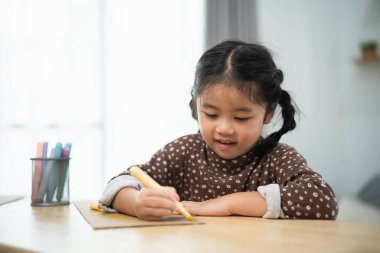 Asyalı genç bir kız elinde sarı bir kalem ve bir kağıt parçasıyla bir masada oturuyor. Bir resim çiziyor ve gülümsüyor.