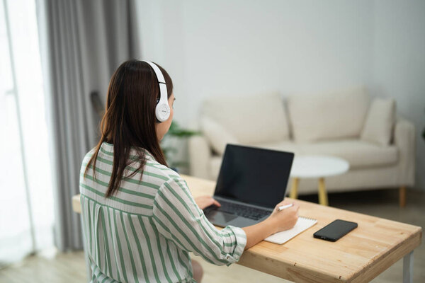 Asian woman is sitting at a desk with a laptop and a notebook. She is wearing headphones and she is working on a project. The room has a couch and a potted plant in the background