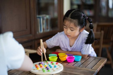 Asyalı genç kız renkli topları olan bir oyunla oynuyor. Topları toplamak için çubuk kullanıyor. Bir kadın onu izliyor.