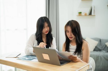 İki Asyalı kadın önlerinde bir laptop ve kağıtlarla bir masada oturuyorlar. Önemli bir şeyi tartışıyorlar.