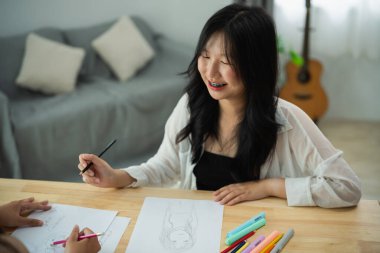 Asyalı bir kadın bir kişinin çizimiyle masada oturuyor. O gülümsüyor ve bir kalem tutuyor. Sahne, kadının çizim sürecinden zevk aldığı yaratıcı ve olumlu bir atmosferi işaret ediyor.