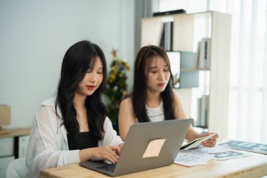 İki Asyalı kadın bir dizüstü bilgisayar ve bir yığın kağıtla bir masada oturuyorlar. Birlikte bir proje üzerinde çalışıyorlar.