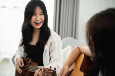 Başka bir kız onu izlerken Asyalı genç bir kadın gitar çalıyor. Ukulele çalan kız gülümsüyor ve diğer kız da oturma odasında gösteriden zevk alıyor gibi görünüyor..
