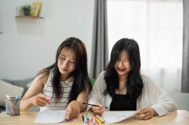 Masada oturmuş yazı yazan iki Asyalı kadın. İçlerinden biri siyah bir gömlek giyiyor. Masa tahtadan yapılmıştır.