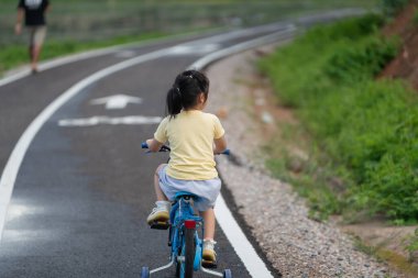 Asyalı genç bir kız yolda mavi bir bisiklet sürüyor. Yol çimlerle kaplı ve arkasında yürüyen bir adam var. Huzurlu ve sakin bir sahne. Kızın keyfi yerinde.
