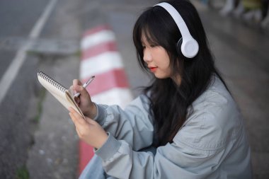 Genç bir kadın, kulaklıkla müzik dinlerken, kaldırımda otururken ve defterine canlı kentsel çevresiyle resim çizerken yaratıcılıktan zevk alır..