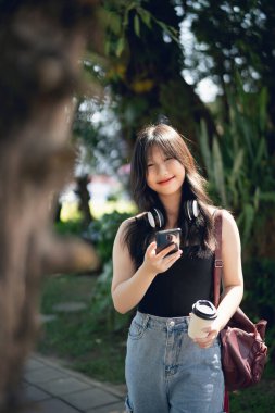 Neşeli genç bir kadın, güneşli bir günün tadını çıkarıyor. Kulaklıkla giyinmiş, akıllı telefonunu ve kahvesini tutuyor. Rahatlama ve mutluluk saçıyor..