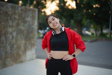 Genç bir kadın spor aletleri ve kulaklıklar takarak, güzel bir günbatımı ortamında pozitiflik ve motivasyon yayarak spora bağlılığını gösteriyor..