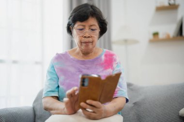 Gözlüklü yaşlı Asyalı kadın evde rahat ve aydınlık oturma odasında, perdeleri ve yumuşak yastıkları olan koltukta otururken akıllı telefon okuyor.