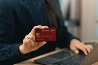 Bilgisayarın başında otururken kredi kartı tutan kişi, ev ofis ortamında online ödemeler ve modern dijital işlemler kavramını vurguluyor.