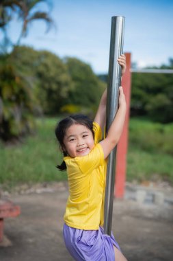 Neşeli Asyalı çocuk canlı bir oyun bahçesinde parlak bir metal direkte oynuyor, güneşli bir ortamda mutluluk ve hayat enerjisini gösteriyor.