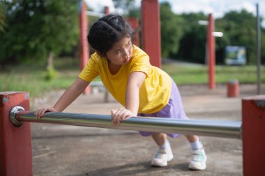 Güneşli bir parkta, oyun parkı ekipmanları üzerinde antrenman yapan Asyalı genç kız spor ve açık hava aktivitelerine azim ve neşeyle odaklanıyor..