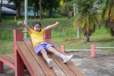 Mutlu Asyalı çocuk güneşli bir günün tadını çıkarıyor. Ağaçlarla dolu yeşil bir parktaki ahşap bir oyun parkında kayarak neşeli anlar için renkli çevreler çiziyor.