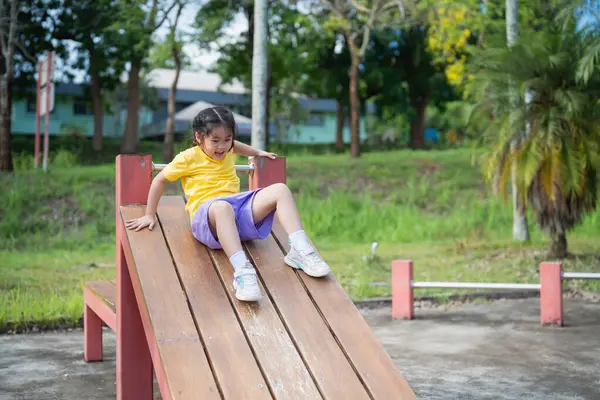 Asyalı küçük bir çocuk oyun alanında güneşli bir günün tadını çıkarıyor. Tahta bir oyun yapısından kayıyor. Kahkahalar ve neşe açık havada oyun zamanı eğlencesine yakalanıyor.