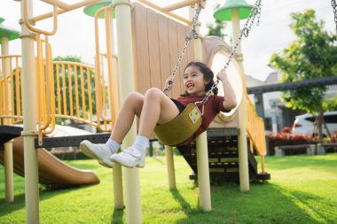 Güneşli bir parktaki salıncakta neşeyle sallanan genç bir kız, yeşil ve parlak ekipmanlarla çevrili dışarıda oyun saatinin tadını çıkarıyor.