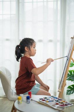 Evde resim yapan, suluboya resim yapmaya odaklanan, güneşli bir pencerenin kenarında oturan yaratıcı ifade, çocukluk sevinci