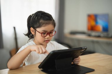 Gözlüklü genç kız parlak odada tablet kullanıyor, dijital dünyayı ve teknolojiyi keşfediyor, evde öğrenme ve yaratıcılıkla meşgul.
