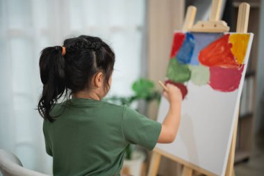 Genç bir kız, parlak odada boya fırçasıyla tuval üzerinde renkli sanat eserleri yaratıyor, yaratıcılığı ve hayal gücünü sanat ifadesiyle keşfediyor.