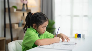 Küçük bir çocuk evde yaratıcı çalışma aktivitesiyle meşgul, renkli malzemelerle parlak, rahat kapalı bir ortamda yazma ve öğrenmeye odaklanmış.