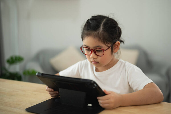 Young girl wearing glasses, focused on tablet screen while sitting at a wooden table in a bright, modern indoor setting with soft furnishings