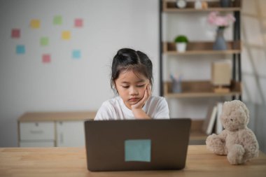 Yumuşak oyuncaklar ve duvarında renkli yapışkan notlar olan rahat bir odada, ahşap masada dizüstü bilgisayar kullanan düşünceli bir kız..