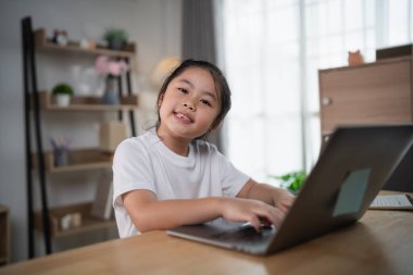 Keyifli genç kız internette öğrenim görürken rahat ev ofisinde dizüstü bilgisayar kullanıyor, modern eğitim ortamı ve teknoloji kullanımı sergiliyor.