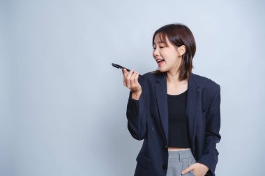 Büyük boy siyah ceketli genç bir kadın açık mavi arka planda ses kaydedicisine gülerek sohbete neşe ve coşku katıyor.