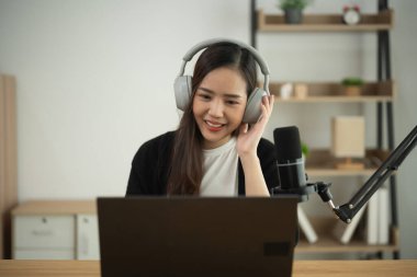 Genç bayan içerik yaratıcısı kulaklık takarak gülümserken mikrofon ve dizüstü bilgisayarla modern çalışma alanında podcast kaydediyor.