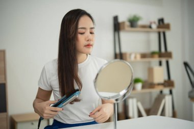 Genç bir kadın, modern ev ortamında düz ütüyle uzun saçlarını şekillendiriyor, özel bir olaya ya da parlak iç mekanlara her gün bakmaya hazırlanıyor.