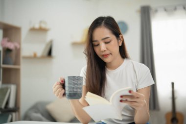 Rahat bir oturma odasında kahvesini yudumlarken, rahat bir anın tadını çıkaran genç bir kadın, yumuşak doğal ışıkla evde dinleniyor.