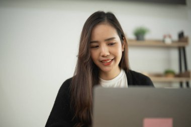 Genç bir kadın modern çalışma alanında çalışırken gülümsüyor yumuşak doğal ışık altında dizüstü bilgisayar kullanıyor, pozitif çalışma ortamını teşvik ediyor ve üretkenliği artırıyor